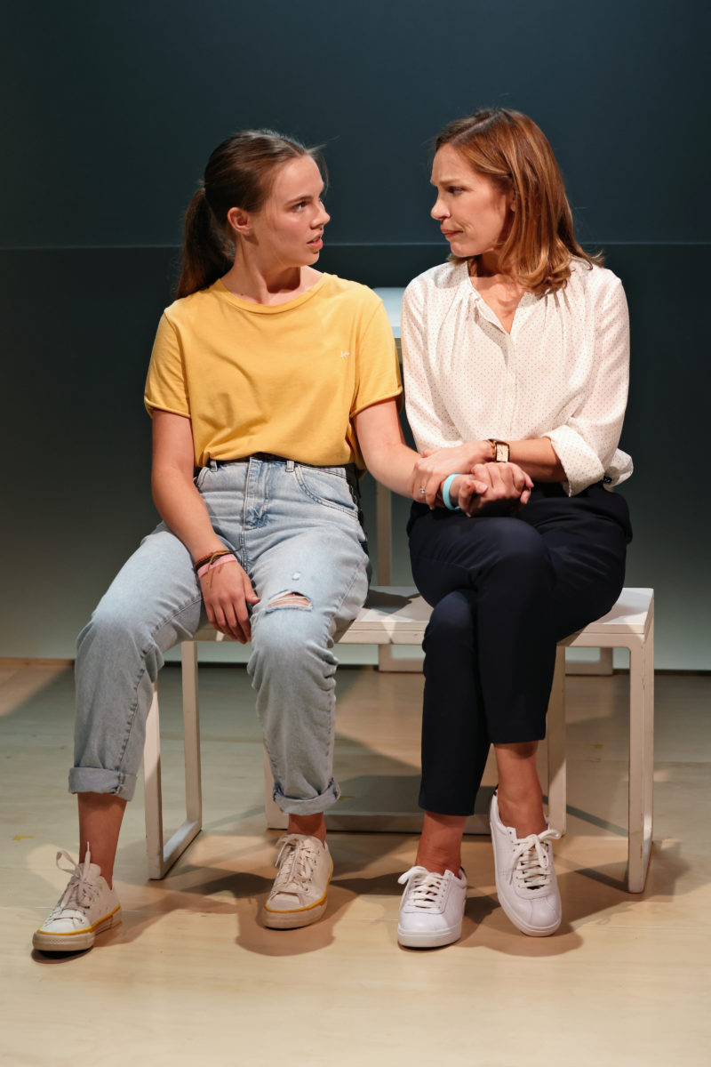 Two women sit closely on a bench, holding hands and looking at each other seriously. One wears a yellow t-shirt and jeans, the other a white blouse and navy trousers. The scene appears calm but emotionally charged.