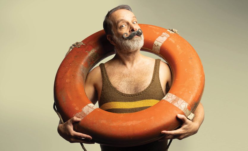 A man with a large moustache and beard, wearing a vintage brown striped swimming costume, stands inside an orange lifebuoy and looks upwards with a playful expression against a plain background.