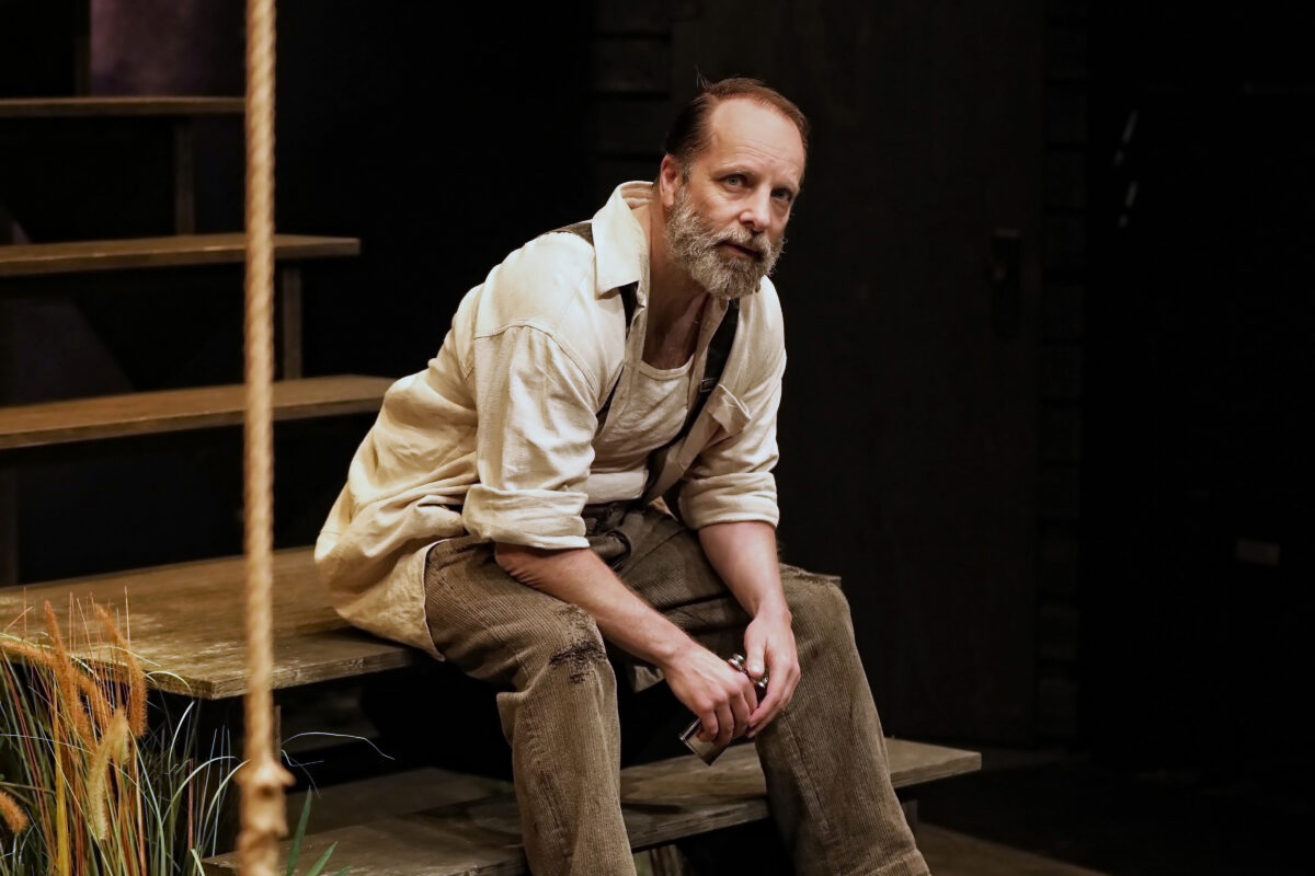 A bearded man wearing a light shirt and brown trousers sits on wooden steps, looking thoughtful. The background is dark with some plants visible in the foreground.