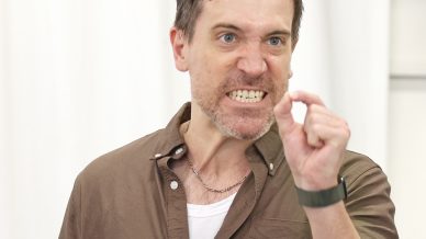 A man with an angry expression holds a wine glass in one hand and gestures with the other, pinching his fingers together as if emphasising a small amount. He is wearing a brown shirt and standing in front of a white background.