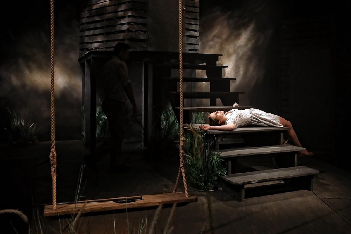 A woman in a white dress lies on wooden steps, dramatically lit on a dim stage. A man stands in shadow nearby. A large wooden swing hangs in the foreground, adding to the theatrical, moody atmosphere.