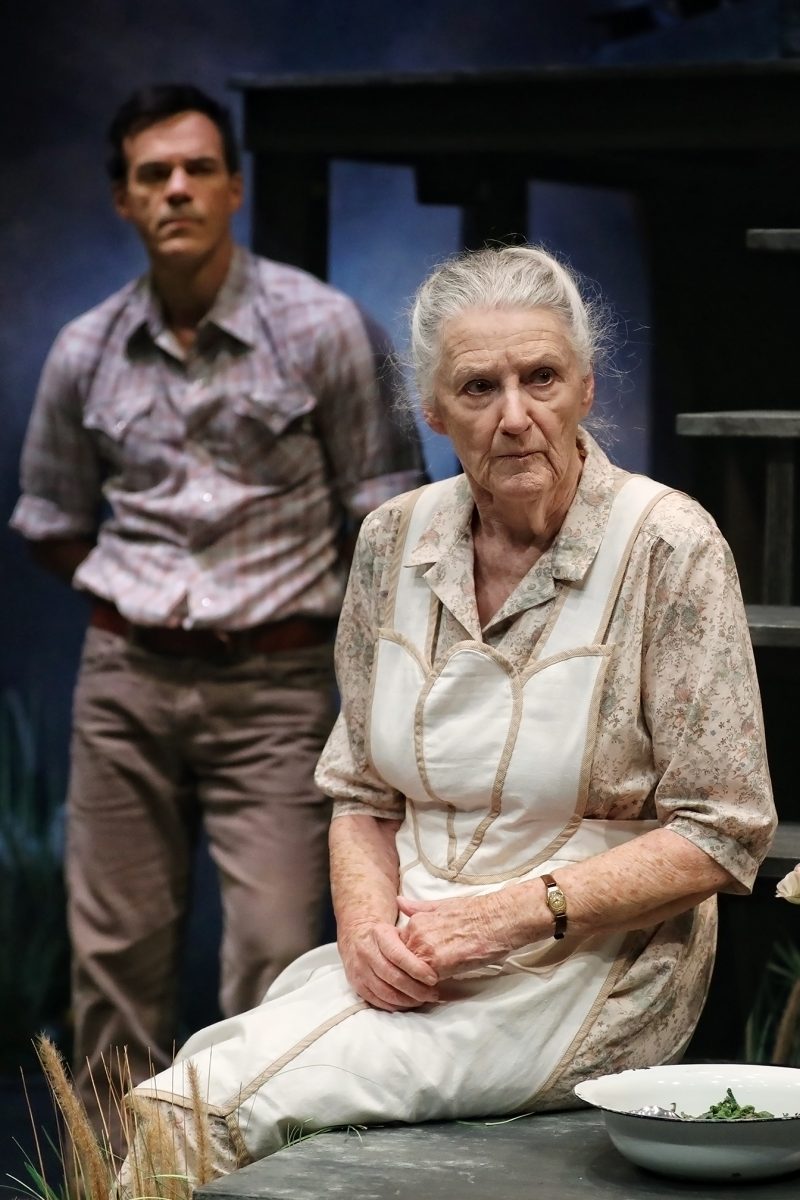 An elderly woman in a floral dress and apron sits with her hands clasped, looking serious. A man in a checked shirt stands behind her, slightly out of focus. The setting suggests a dramatic stage performance.