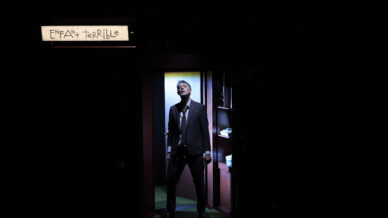 A man in a suit stands dramatically in a doorway, lit by a spotlight, with a sign above reading ENFANT TERRIBLE in handwritten letters. The surrounding area is mostly dark.