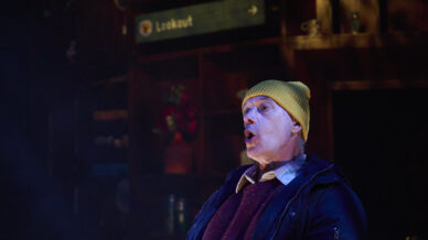 A man wearing a yellow beanie and dark jacket appears to be speaking or singing with an expressive face. Behind him, shelves and a sign that reads Lookout are visible in a dimly lit setting.
