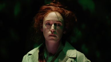 A woman with pale skin and messy red hair stands in dramatic green lighting, looking slightly to the side with a serious expression, wearing a light grey coat over dark clothes.