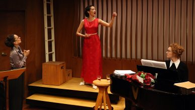A woman in a red dress passionately sings on a small stage. Another woman stands nearby, watching her, whilst a third woman sits at a piano with sheet music, looking up. A glass of water and a bouquet of flowers rest on the piano.