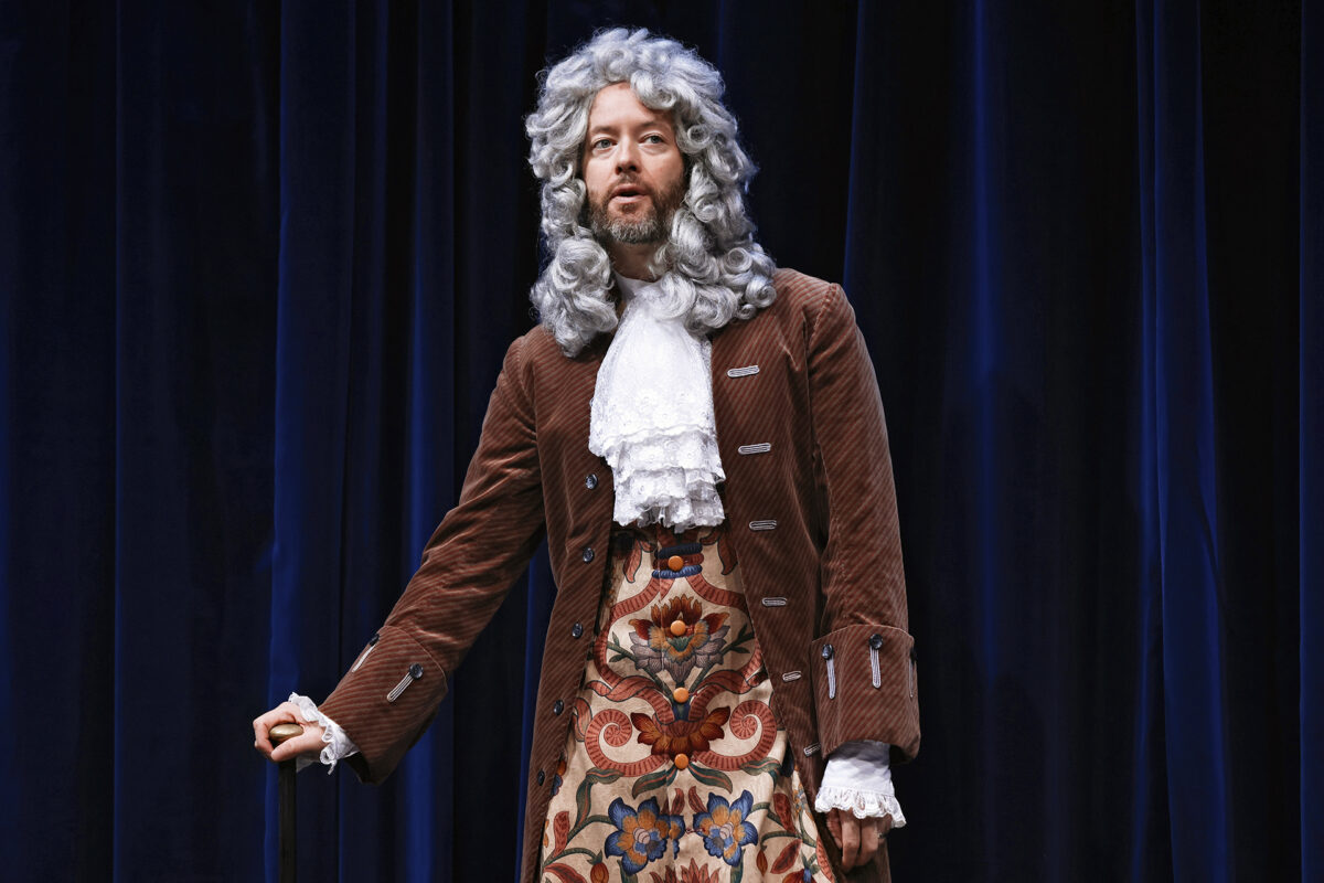 A person in a curly grey wig, brown jacket with silver buttons, white lace collar, and a floral-patterned waistcoat stands against a dark blue curtain, holding a walking stick.