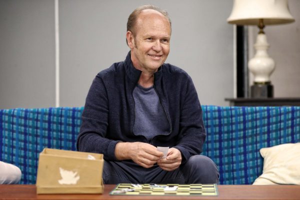 A middle-aged man with short hair sits on a blue checked sofa, smiling and holding a piece of paper. A box and a game board are on the wooden coffee table in front of him, and a lamp is in the background.
