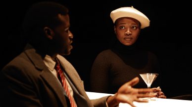 A woman in a white beret and black polo neck sits at a table with a martini glass, listening to a man in a suit and red-striped tie who gestures as he speaks. The scene is dimly lit.