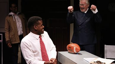 An older man in a suit enthusiastically gestures with raised fists while standing next to a seated younger man in a shirt and red striped tie, who is smiling. A football and papers are on the desk between them. Another person stands in the background.