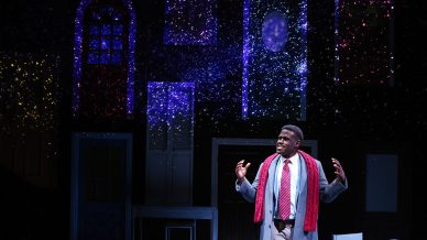 A man in a suit and red scarf stands on stage with hands raised, appearing to speak or perform. Colourful lights and star-like sparkles illuminate door and window frames behind him, creating a magical night-time atmosphere.