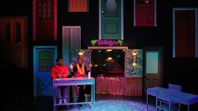 Two people sit at a bar counter under a neon Wallys sign, with colourful doors adorning the dark wall behind them. The stage is lit with vibrant, multicoloured lights. Empty tables are nearby.
