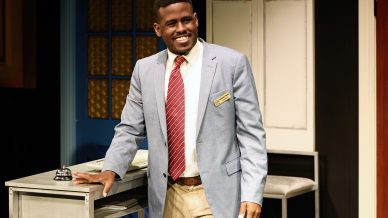 A smiling man in a light blue blazer, red striped tie, and name badge stands beside a desk in an office setting with a bell and papers, against a backdrop of blue and white doors and cupboards.