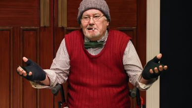 An older man in a wheelchair wearing a grey knitted hat, glasses, a red jumper vest, bow tie, checked shirt, and fingerless gloves gestures with open hands in front of wooden doors.