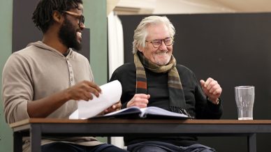Two men sit at a table, smiling and laughing. One holds a script, the other wears glasses and a striped scarf. An empty glass and open book are on the table in front of them.