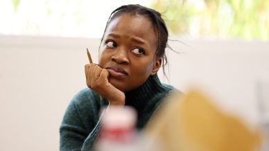 A woman wearing a green jumper rests her chin on her hand and looks up thoughtfully whilst holding a pencil. The background is softly blurred with green foliage and indoor light.