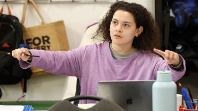 A person with curly hair in a purple shirt gestures with both hands while sitting at a desk with a laptop, water bottle, and papers. In the background are rucksacks and a tan tote bag.
