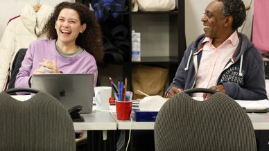 Two people sit at a table, smiling and laughing together. The woman on the left wears a purple shirt, and the man on the right wears a pink shirt with a navy hoodie. Office supplies and a laptop are on the table in front of them.