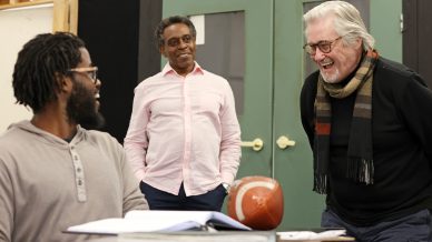 Three men interact cheerfully in a room; one sits at a desk with a football and open book, while the other two stand nearby, smiling and laughing, suggesting a friendly, informal setting.