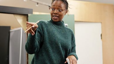 A person wearing glasses and a green jumper gestures with a pencil in one hand and holds a small notepad in the other, appearing to speak or give instructions in an indoor setting.