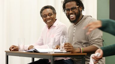 Two men sit side by side at a table, smiling and holding clear glasses. Papers and notebooks are on the table. They appear to be engaged in a lively conversation or discussion. A third person’s arm is visible in the foreground.