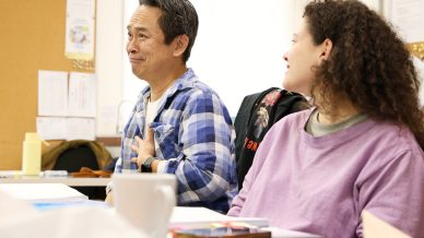 A man in a blue checked shirt sits with his hand on his chest, appearing thoughtful or moved, next to a smiling woman in a lavender jumper. Papers, a mug, and books are on the table in front of them.