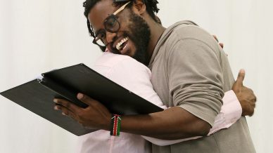 Two people embrace warmly while standing, one holding a black folder and smiling. Both are dressed casually and appear happy, with a light-coloured, plain background behind them.