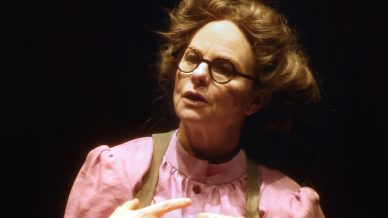 A person wearing round glasses, a pink blouse with puffed sleeves, and a dark waistcoat gestures with their hands while speaking, standing against a dark background.