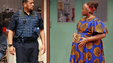 A police officer in uniform stands facing a pregnant woman wearing a colourful, patterned dress and headwrap. They appear to be having a serious conversation in a room with a kitchen and a green door in the background.