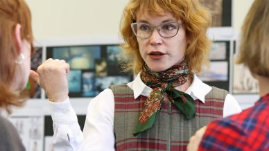 A woman with curly red hair, glasses, and a checked dress gestures whilst speaking to two people, who are seen from behind. Photos and images are displayed on the wall in the background.