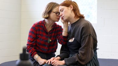 Two women sit close together; one gently touches the other's face, offering comfort. The woman being comforted has her eyes closed and appears sad. They are indoors, near a window, with light coming in.