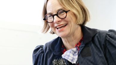 A smiling woman with short, light brown hair and round glasses wears a navy jacket, checked shirt, and a decorative brooch, standing indoors and gesturing with her hands.