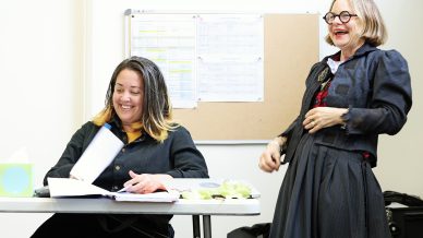 Two women in a bright room; one is sitting at a table looking through a book and smiling, while the other stands nearby, laughing. A noticeboard with papers is on the wall behind them.