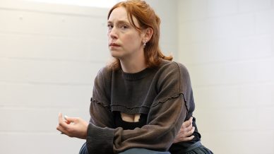 A woman with red hair wearing a dark jumper and skirt sits indoors against a white wall, looking slightly to the side with a thoughtful expression, one arm crossed over her body and holding something small in her hand.