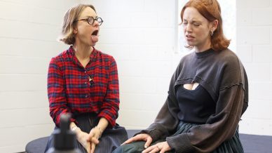 Two women sit indoors; one in a red checked shirt sticks out her tongue and pulls a silly face, while the other in a dark jumper and skirt looks upset or distressed, with her eyes closed.