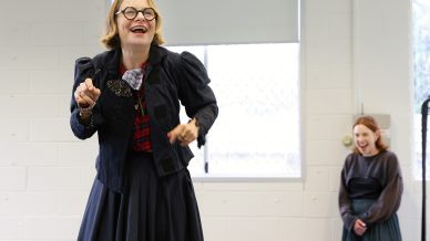 A woman wearing glasses, a black jacket, and a checked shirt smiles and gestures energetically, while another woman in the background stands laughing near a white brick wall and window.