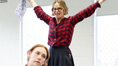 Two women in costume are on a round stage. One sits in front, looking thoughtful, whilst the other stands behind her with arms raised, smiling. They are indoors, with light coming through windows behind them.
