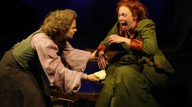 Two women in old-fashioned costumes are on stage; one woman is handing a letter to the other, who appears distressed and is crying out, her face contorted with emotion. The lighting is dramatic and theatrical.