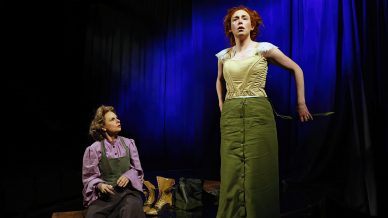 Two women in period costumes are on stage; one sits and looks up whilst the other, with red hair, stands adjusting her green skirt and corset against a dramatic blue curtain backdrop.