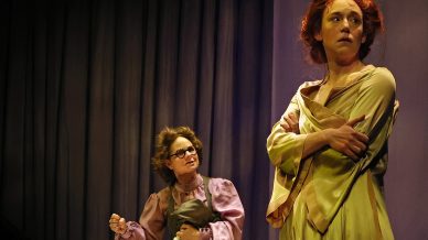 Two actors perform on stage; one wears a long light green dress and stands barefoot, arms crossed and looking to the side. The other, in glasses and a purple blouse, clutches clothing. Period boots are on the floor in front.