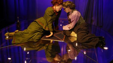 Two women in period costumes kneel closely on a glass stage, leaning in towards each other over a board game, under dramatic purple lighting with their reflections visible below.