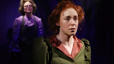 Two women in period costumes stand under dramatic stage lighting; the woman in front looks distressed, with reddish hair and a green dress, while the woman behind her wears glasses and a purple blouse, both against a dark background.