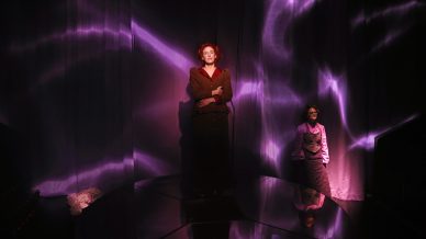 Two people stand on a dimly lit stage with purple, wavy light patterns projected on dark curtains behind them. One person stands at the centre in a brown coat, while the other stands to the right in a purple outfit.