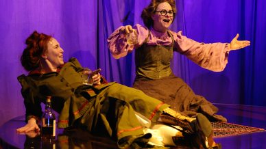 Two women in vintage costumes laugh and hold drinks onstage; one reclines on a table with a bottle beside her, while the other sits with arms spread wide. Purple stage lighting and curtains create a theatrical atmosphere.
