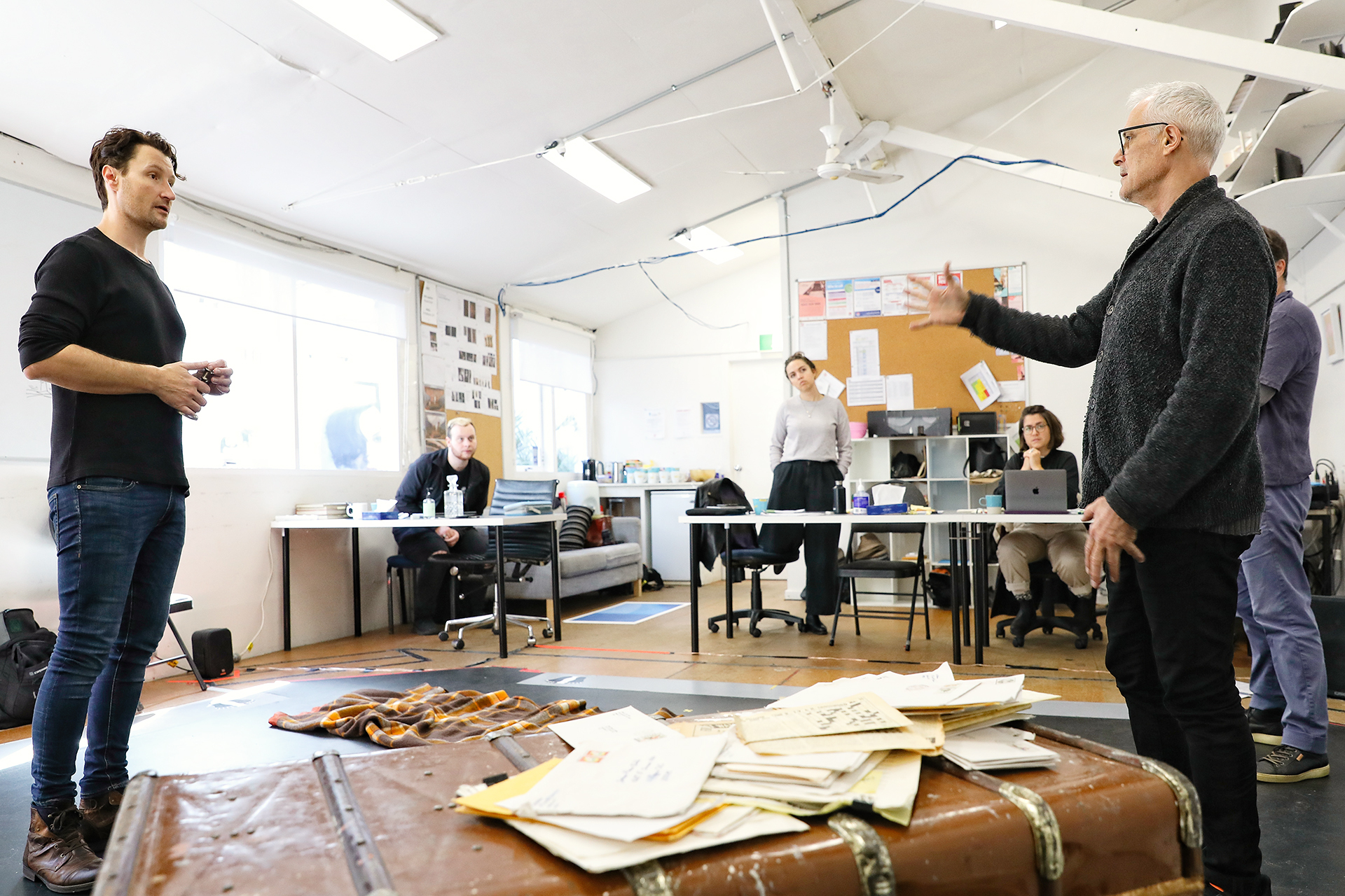 A group of people in a brightly lit room, some sitting at desks with laptops and papers, whilst two people stand facing each other and gesturing, suggesting a collaborative or rehearsal setting.